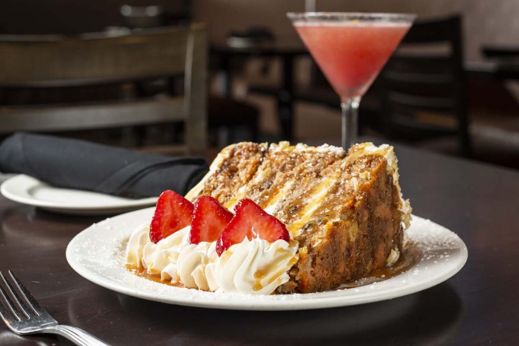 Slice of carrot cake on its side with whipped cream and strawberry garnish and pink martini cocktail in background