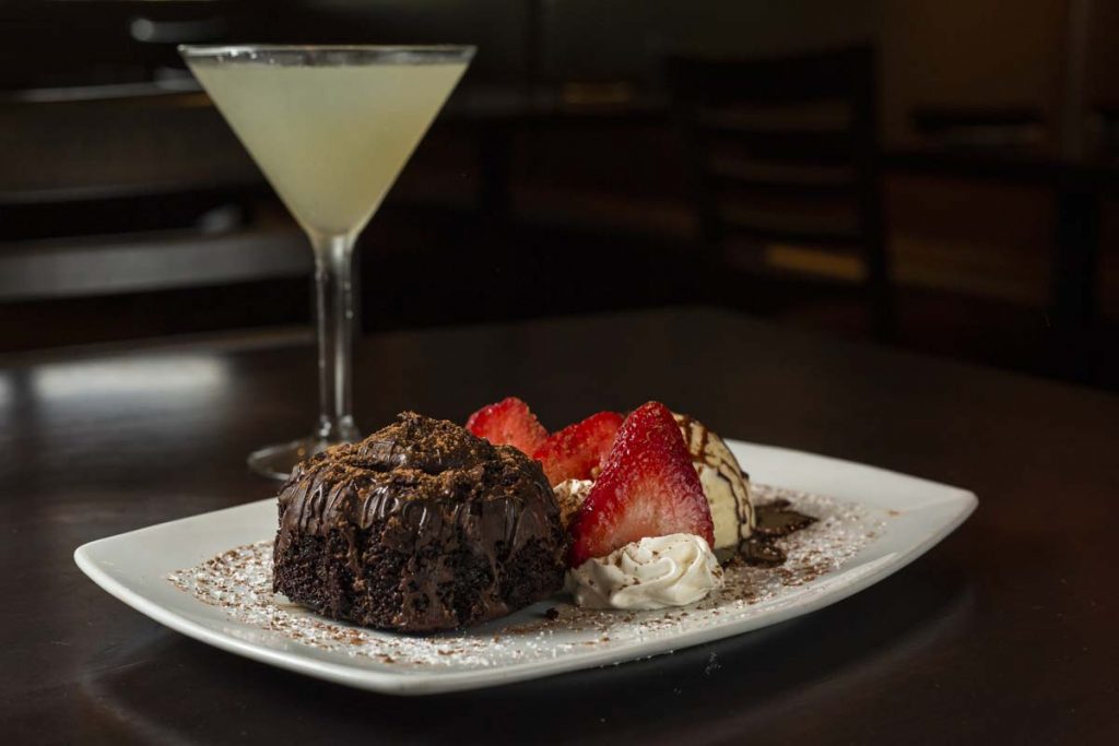 Chocolate lava cake garnished with ice cream and strawberries with lemon martini cocktail in background