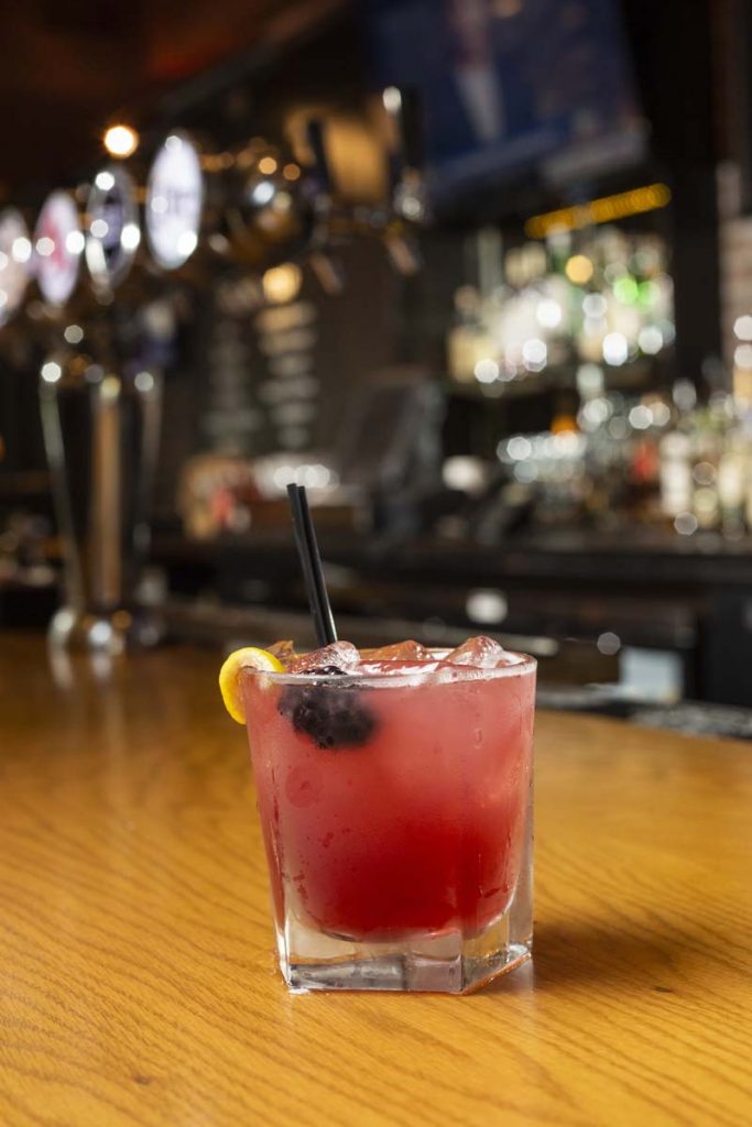 Pink cocktail in rocks glass with lemon and blackberry garnish sitting on oak bar.