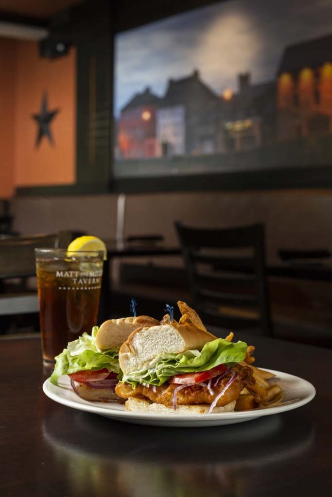 Fried fish sandwich garnished with lettuce, tomato and plated with French fries with glass of iced tea and lemon in background.